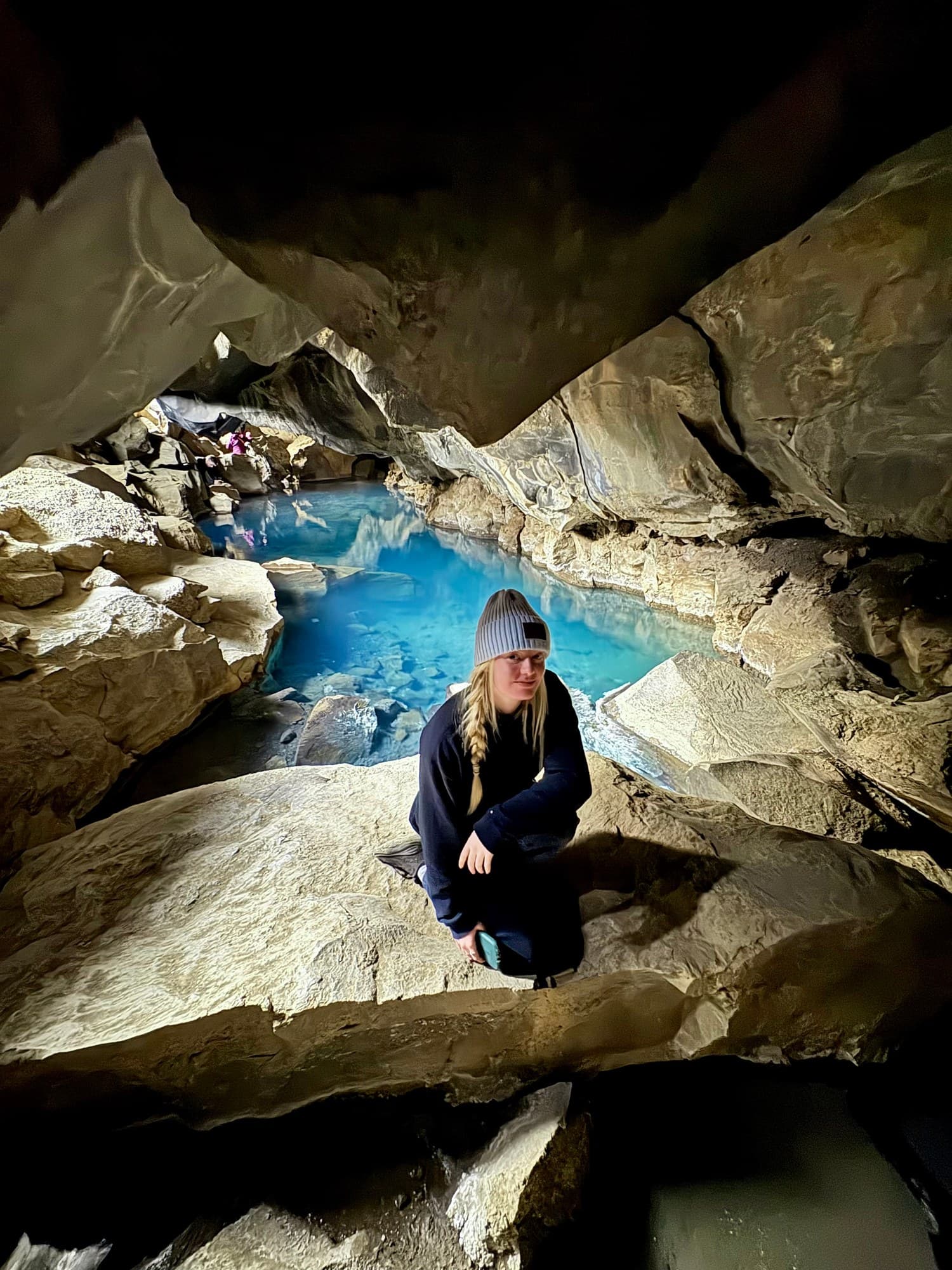 Hannah seated on a dry rock beside the blue pool inside Grjótagjá, with daylight entering behind her.