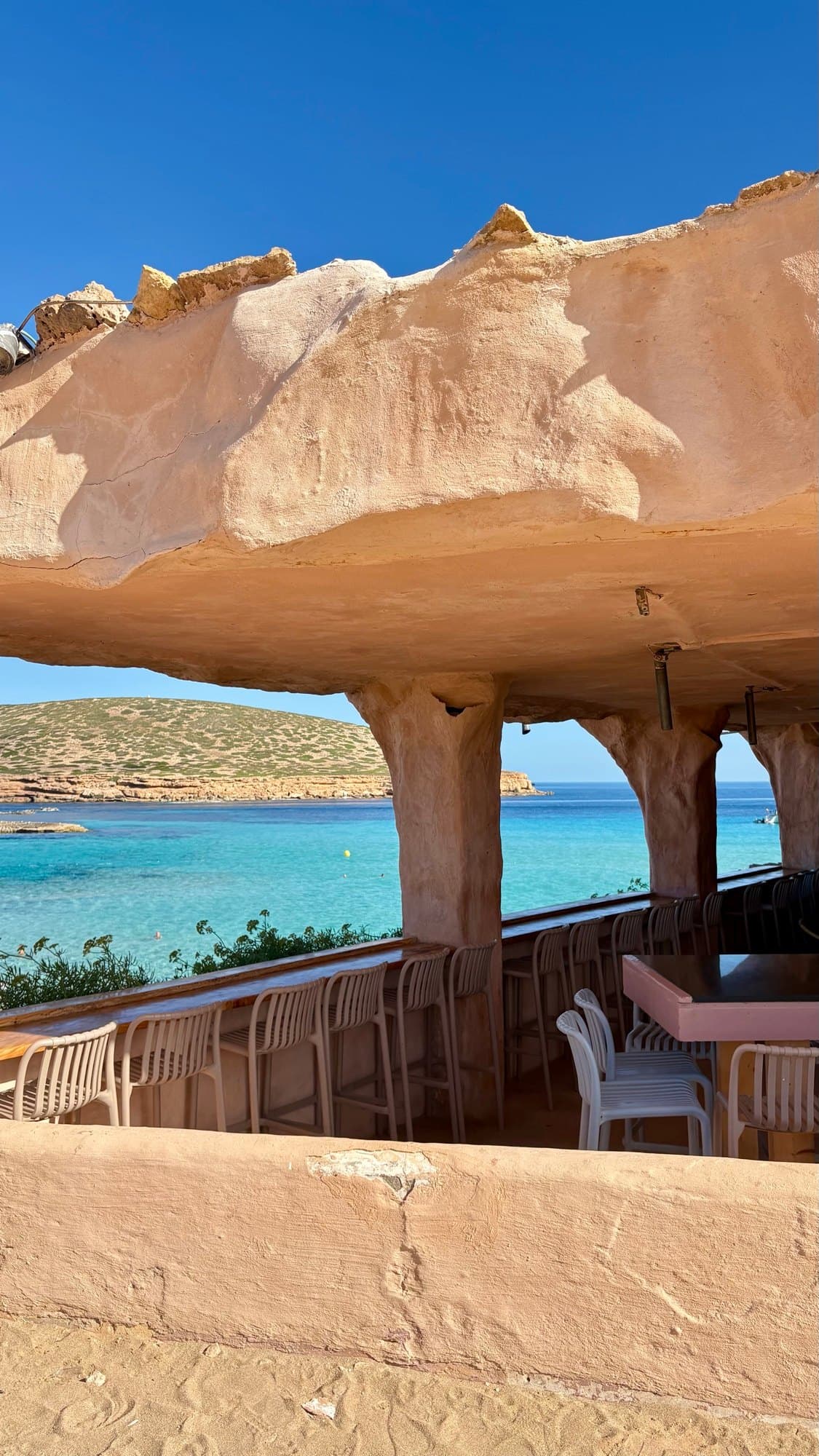 Turquoise sea and a low island seen through the warm stone openings of a shaded terrace in Ibiza.