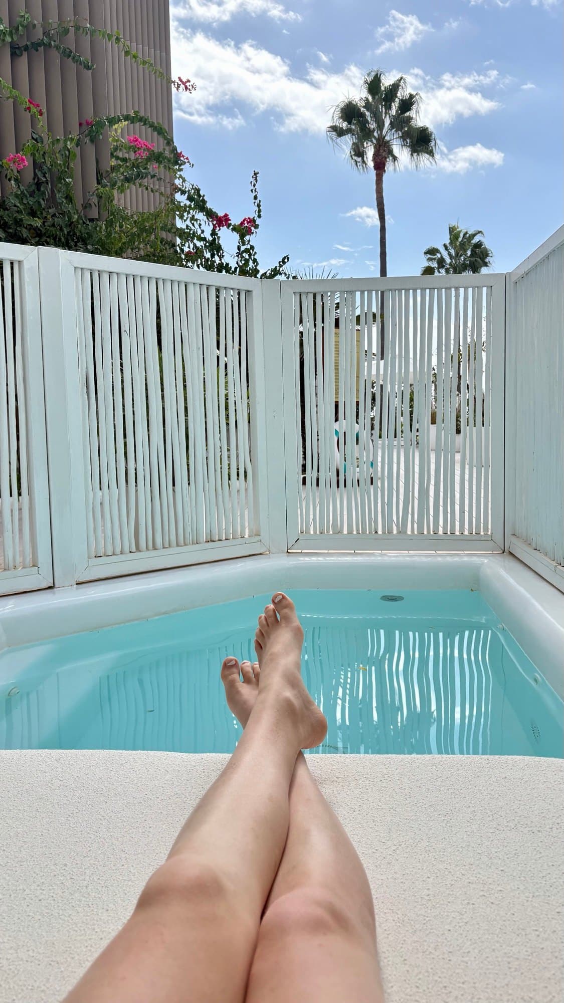 A small turquoise pool behind a white slatted screen, with palms above and a traveler’s legs in the foreground.
